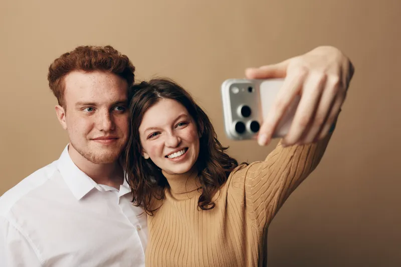 Young couple takes a selfie with a smartphone on beige background. Smiling woman in brown sweater holds phone, man in white shirt stands close. Concept of friendship and technology.