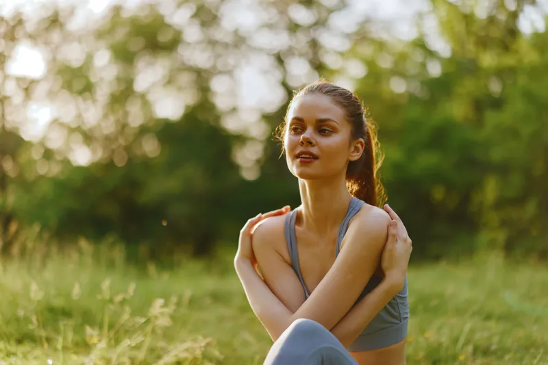 Young athletic woman in casual sportswear sitting in a sunny park, enjoying nature, embodying relaxation and mindfulness in a vibrant green environment
