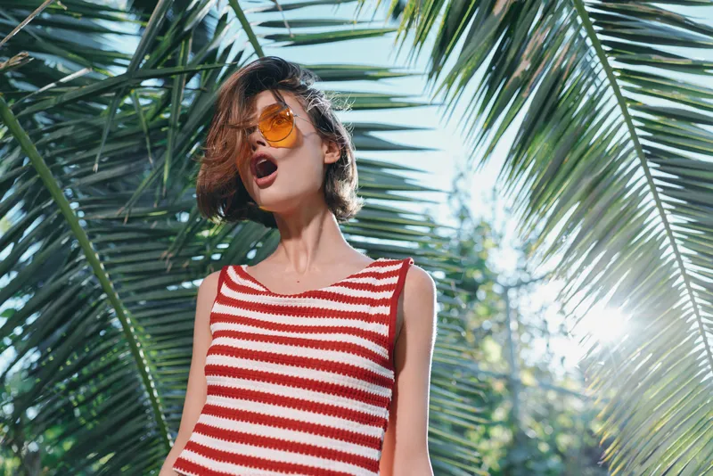 A stylish woman wearing a red and white striped top stands outdoors among lush palm leaves, sunglasses framed by bright sunshine, radiating bold confidence and a sunny, dynamic mood.