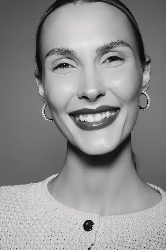 Smiling white woman with light skin and short hair wears earrings and knitted sweater in studio portrait.