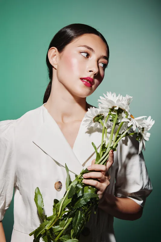 Young woman with glowing skin and glossy makeup holds a bouquet of white flowers over green background. Creative beauty and fashion concept with fresh natural style features.