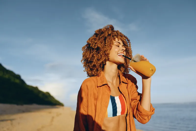 A happy young woman with curly hair drinks orange juice through a straw on a sunny beach. She wears a colorful swimsuit and an orange shirt, enjoying a relaxing summer day outdoors.
