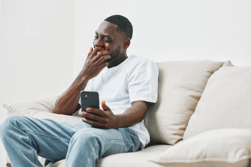 Smiling African American man using smartphone while sitting on couch at home, wearing casual white t shirt and jeans, showcasing joy and relaxation