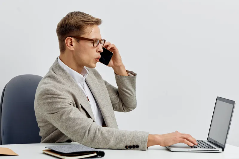 Businessman using laptop and talking on mobile phone while sitting at desk in modern office workspace. Professional, focused, corporate, technology, communication, indoors, modern lifestyle concept, l