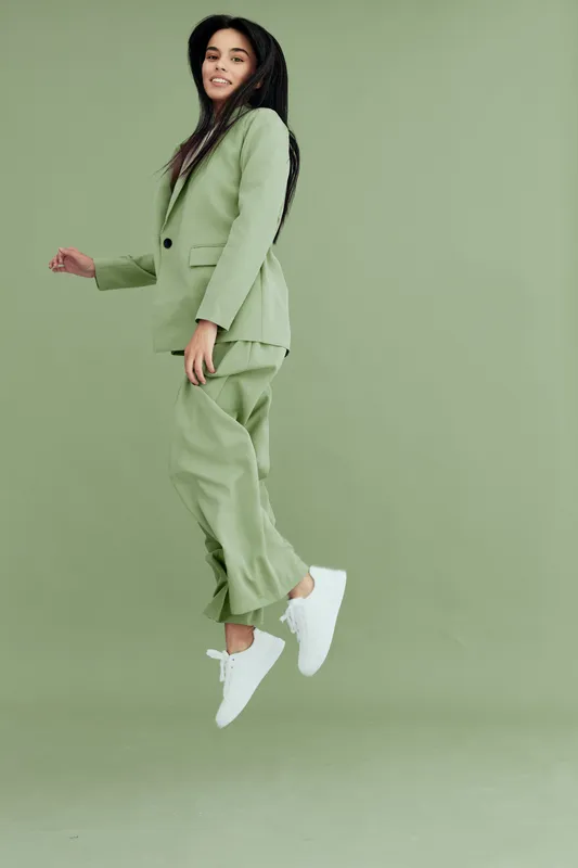 young woman jumping in green suit and white sneakers on green background, smiling happy joyful energetic casual outdoors