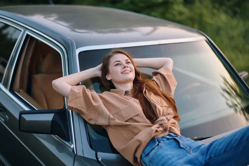 Young woman relaxing on a vintage car, wearing casual beige shirt and blue jeans, smiling contently in natural outdoor setting during sunset, evoking a sense of freedom and joy.