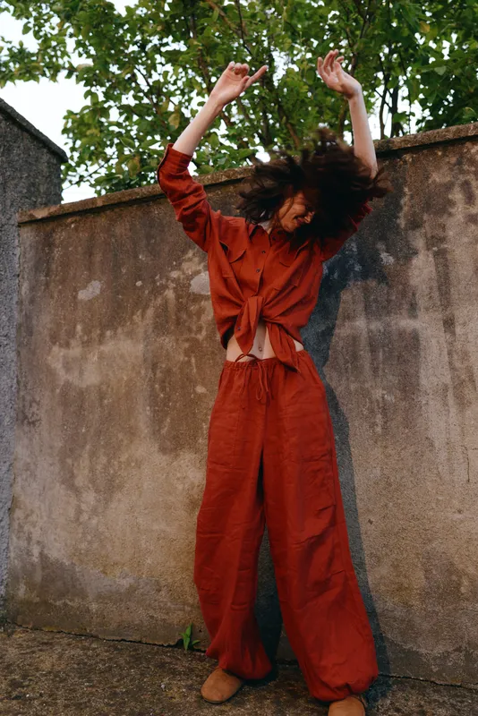 Woman in a red wideleg outfit dances outdoors beside a weathered wall, mid motion pose. Emphasis on lifestyle, movement, fashion, energy and joy