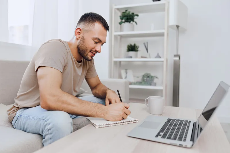 Thoughtful man writing notes in a cozy modern living room, surrounded by greenery and natural light, conveying creativity and focus in a homely atmosphere.