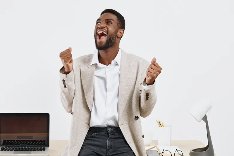 Smiling man with beard, joyful expression, wearing smart casual blazer and white shirt, standing in modern office with laptop, celebrating success, light background, positive work attitude, lifestyle 