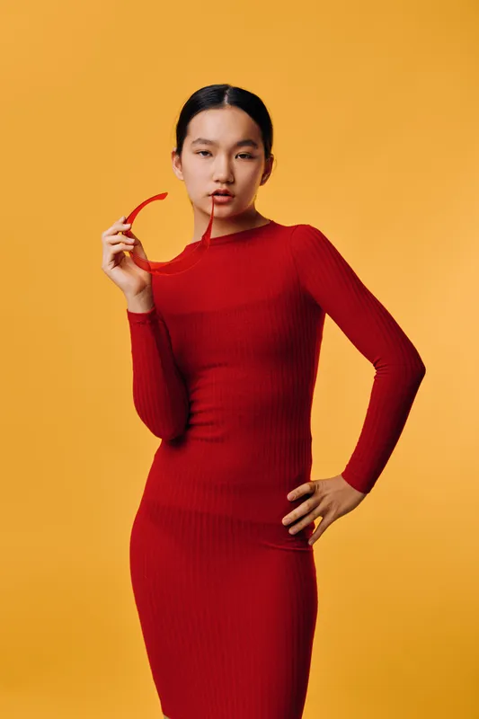 Woman in red dress posing with sunglasses in hand against orange background. Elegant fashion model standing with confident expression and stylish look for portrait or style concept.