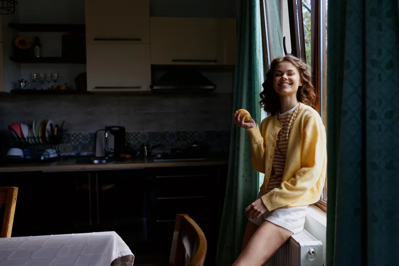 Young woman with curly hair stands by a sunlit window indoors, wearing a yellow cardigan and striped top, smiling softly as warm light fills the cozy kitchen space
