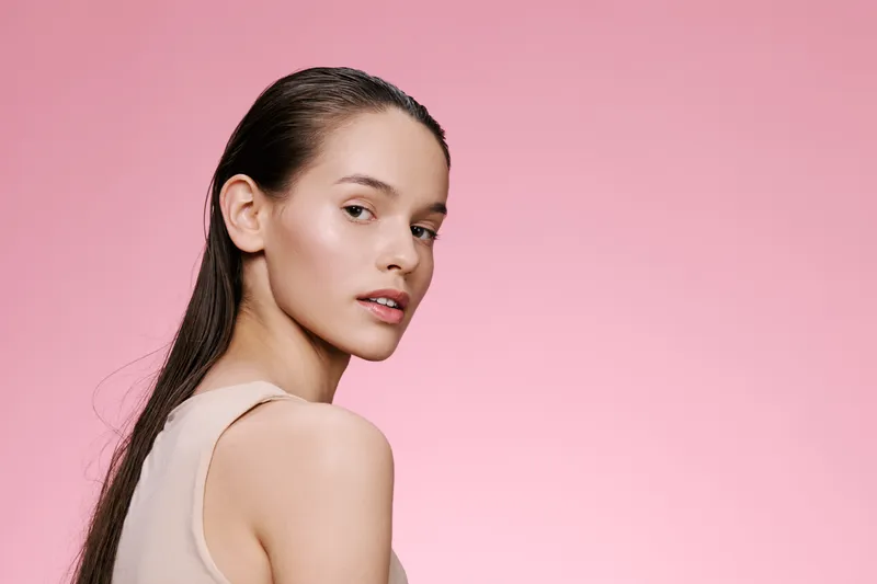 Young woman with long hair looking back, soft lighting, minimalist style, and pastel pink background, represents beauty and self care