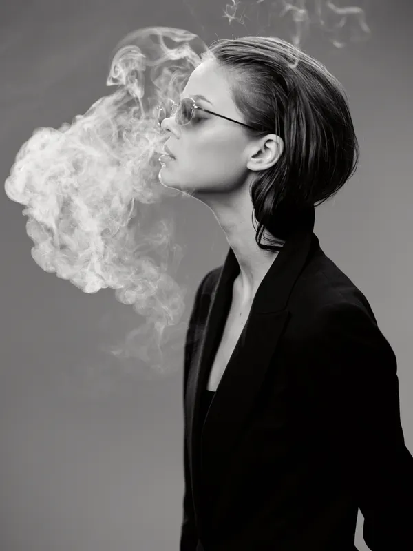 Woman in black blowing smoke wearing sunglasses with slicked-back hair and stylish blazer in black and white