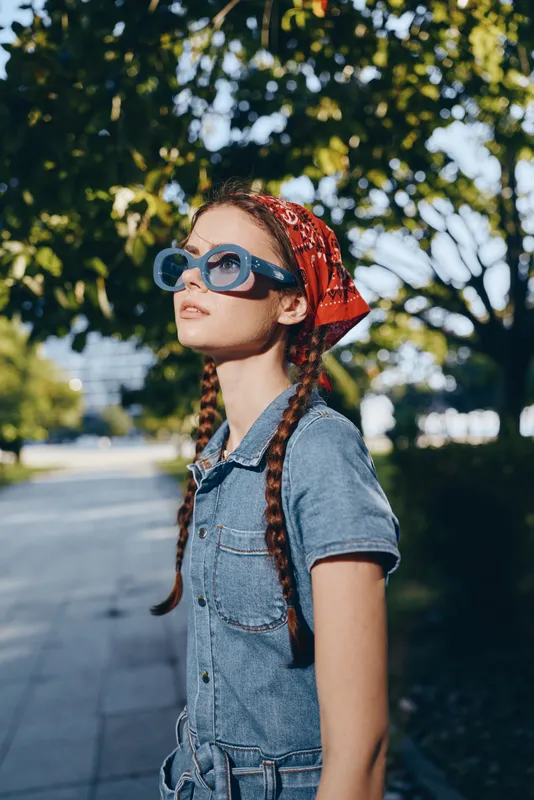 young woman with braided hair wearing denim outfit and red headscarf looking thoughtfully outdoors in sunny urban park setting