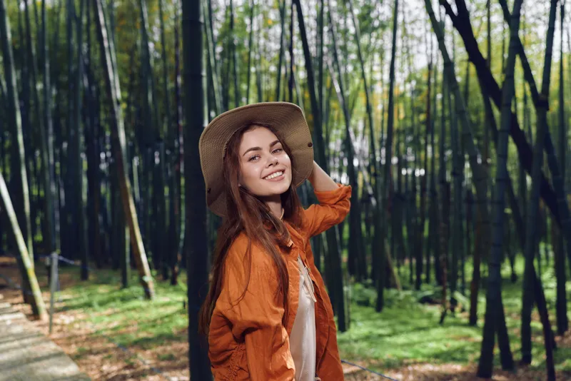 Woman portrait smile in bamboo forest wearing straw hat and orange jacket, nature outdoors scene of happy young traveler enjoying a serene green walkway with soft sunlight.