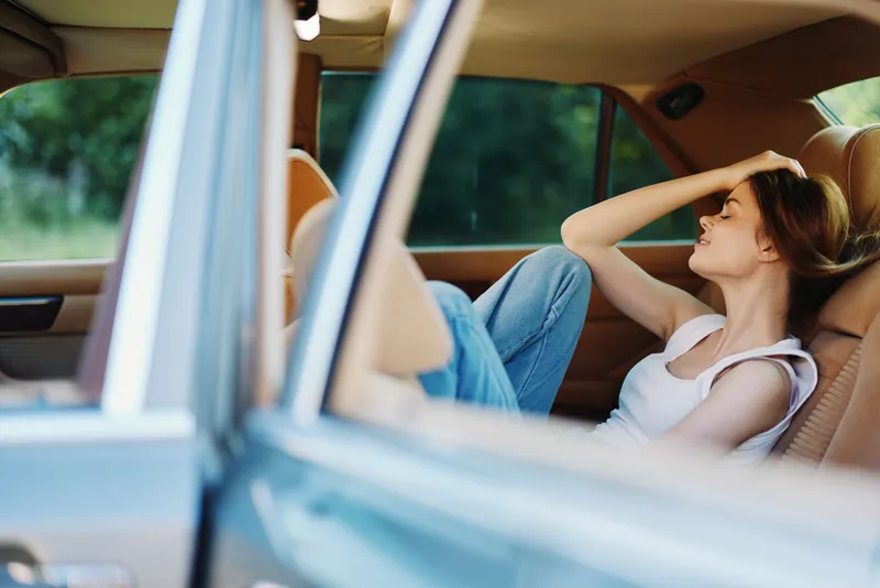 Young woman sitting in a car, expressing feelings of stress and reflection, dressed casually in a tank top and jeans, with a blurred green background.