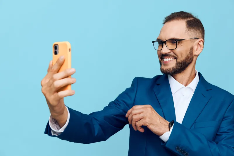 A professional man in a blue suit takes a selfie against a vivid color backdrop, showcasing confidence and joy. The isolated color background highlights his business presence and positive emotion for