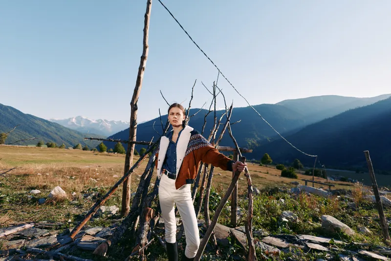 Man portrait in mountains and nature, standing in rural field by rustic fence, fashion jacket and white pants, outdoors lifestyle and scenic landscape with distant hills and clear sky.