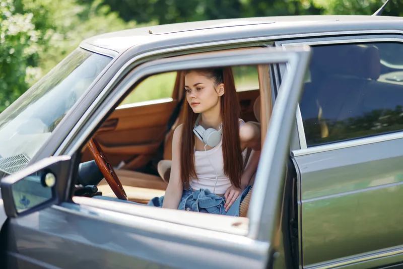 Young woman with long hair in casual attire sitting in a vintage car, enjoying a sunny day outdoors, exuding a relaxed and carefree vibe.