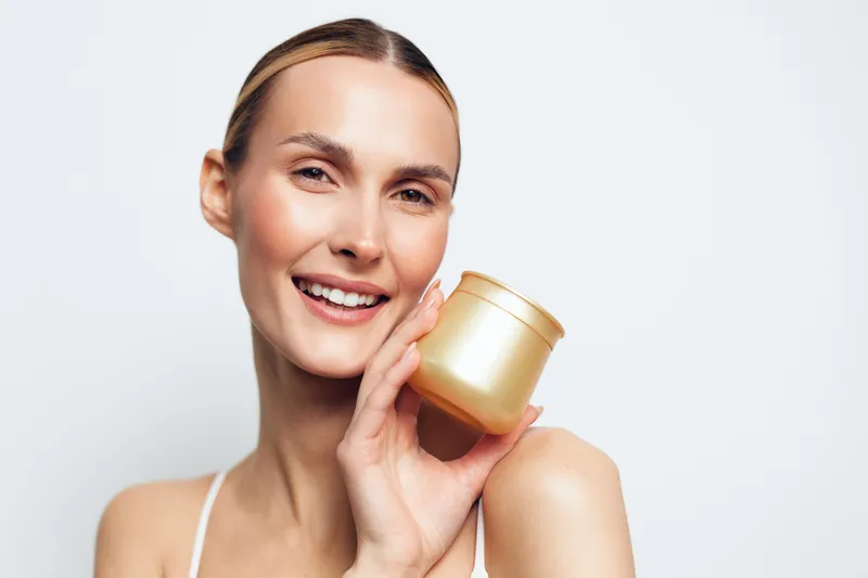 Smiling woman holding golden cosmetic jar near face, clear skin, natural makeup, bright background, skincare routine, beauty concept, fresh and healthy look.