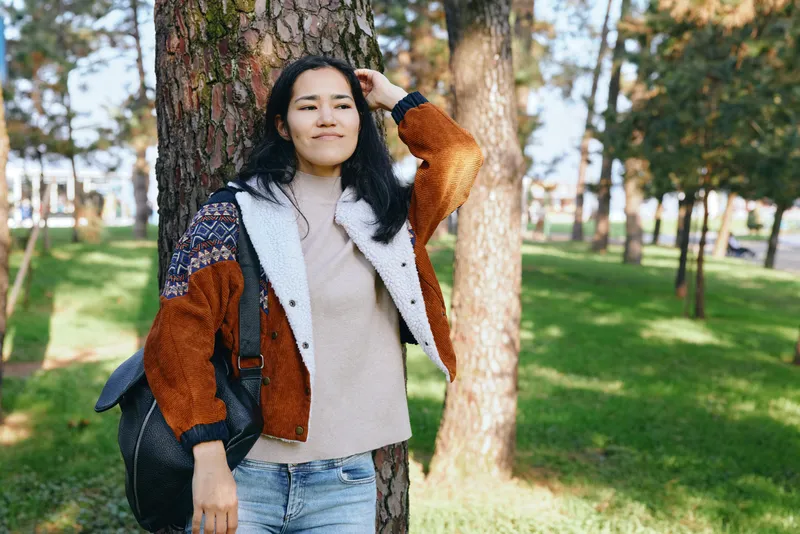 Casual outdoor scene with a woman leaning against a tree in a park, wearing a color block jacket and knit sweater, carrying a backpack. Sunny day, calm mood, nature backdrop and fresh air.