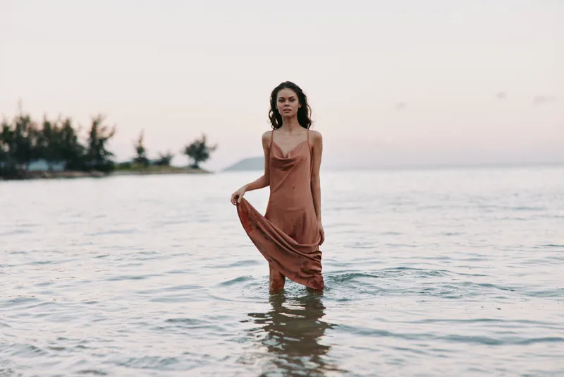 Woman standing in calm sea water at sunset wearing a flowing brown dress with serene expression and soft natural light.