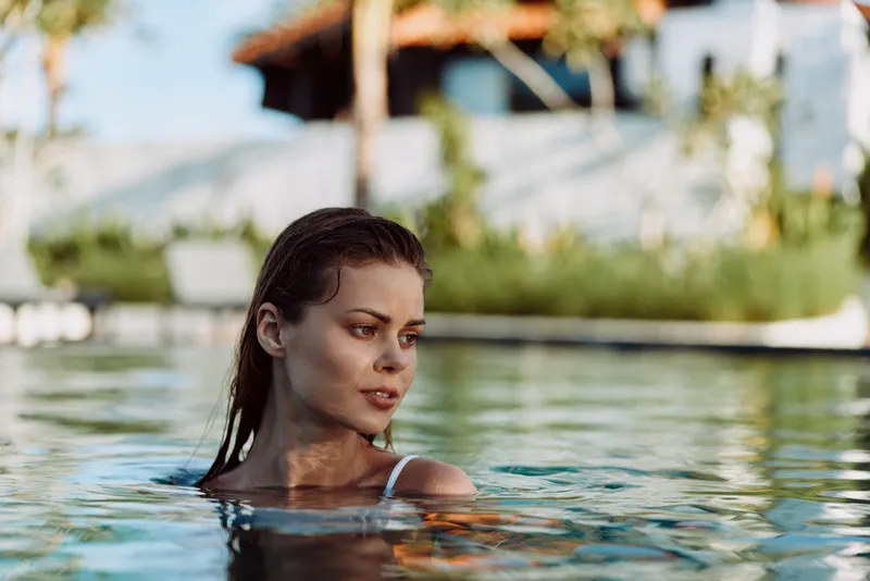 Woman in a swimming pool, gazing quietly as water ripples around her. Sunlit outdoor scene conveys relaxation, vacation vibes, and a calm, reflective mood in a lifestyle setting.