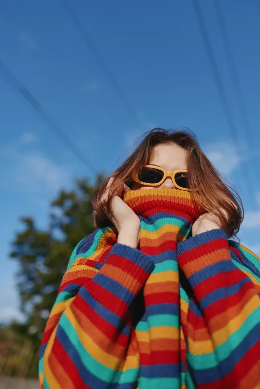 Rainbow sweater striped sweater sunglasses woman portrait outdoor colorful fashion. Young woman pulls oversized striped pullover up over her face, yellow sunglasses, playful pose under blue sky.