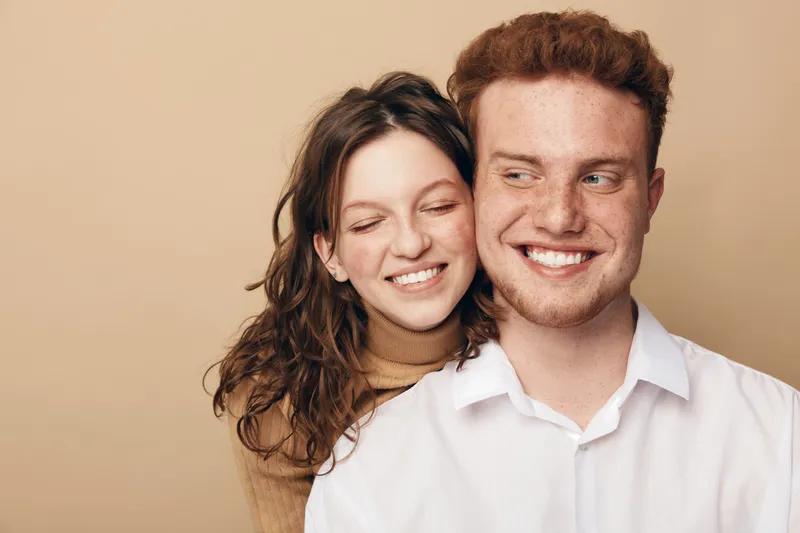 Smiling young couple with curly hair and freckles embraces against beige background. They show affection and happiness, representing love, friendship, and positive emotions in casual style.