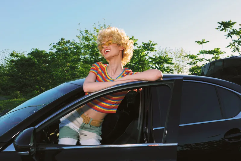 A cheerful woman leans out of a black car window in bright retro film tones, wearing a striped top and denim shorts. A lighthearted lifestyle moment captured in warm, cinematic color.