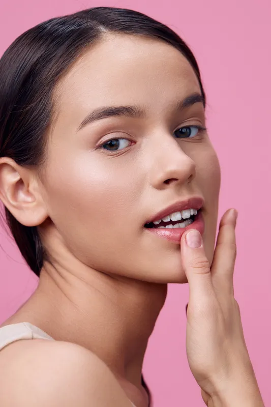Portrait of a young woman touching her face, with glowing skin and a soft expression, showcasing beauty against a pink background