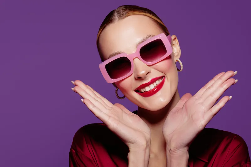 Smiling woman with pink sunglasses and red lipstick poses with hands near face on purple background. Concept of fashion, beauty, style, and confident female model portrait.