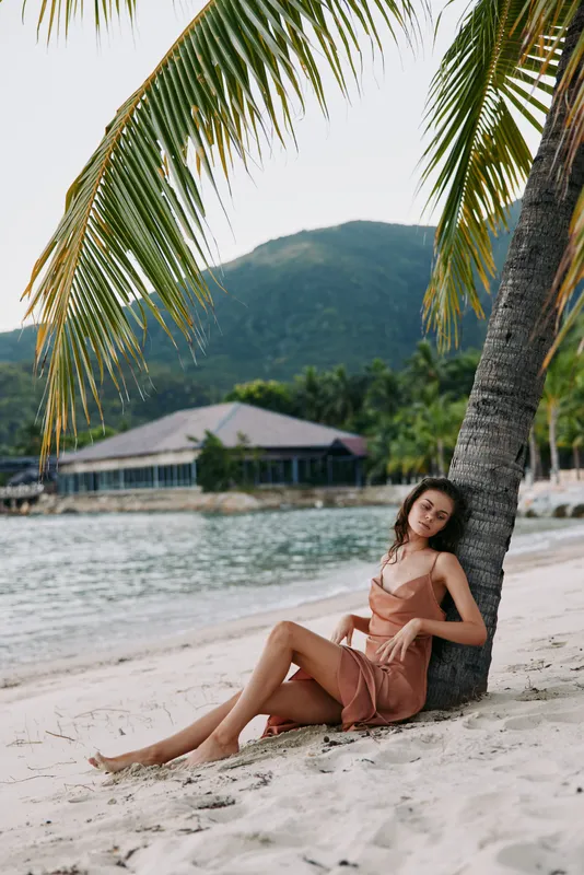 Relaxed woman in summer dress sitting on sandy beach under palm tree with tropical mountains and calm sea in background during sunny day