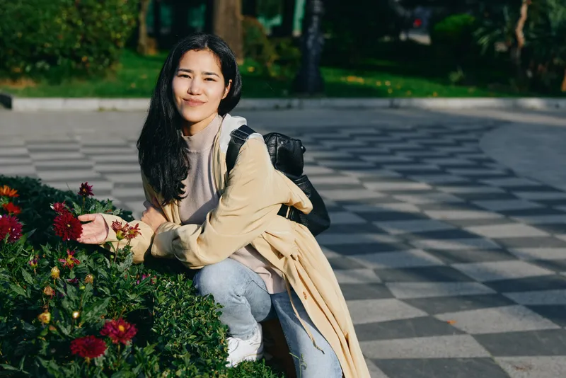 Woman with backpack kneels beside vibrant flowers in a quiet park, enjoying a sunny day, casual sweater and jeans, relaxed mood, candid outdoor photography with natural light and fresh breeze