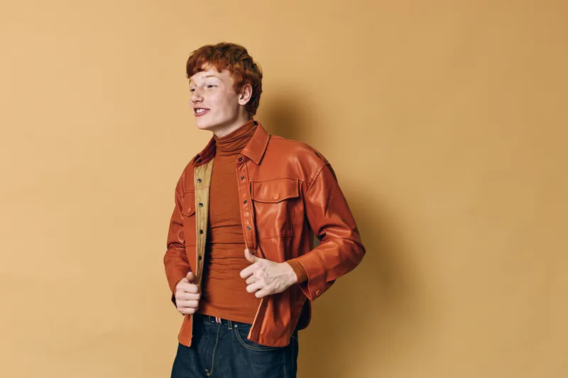 Young man with red hair wearing brown leather jacket and turtleneck posing against beige wall. Casual fashion style, confident expression, trendy outfit and modern look indoors.
