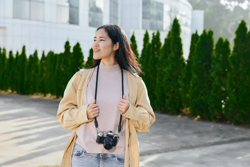 Woman with camera around neck in a beige coat standing outdoors, smiling, bright sunlit day among trees and modern buildings