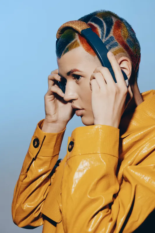 young woman with colorful hair wearing headphones and a yellow jacket, focused expression, modern style, vibrant blue background, creative fashion concept