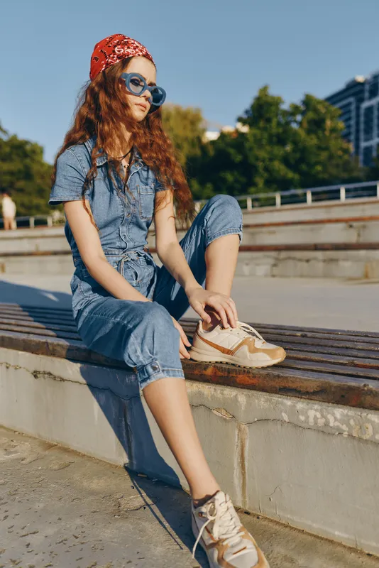 lifestyle woman in modern boho-western denim street style sitting outdoors with sunglasses and bandana, casual urban fashion, film color tones, relaxed pose