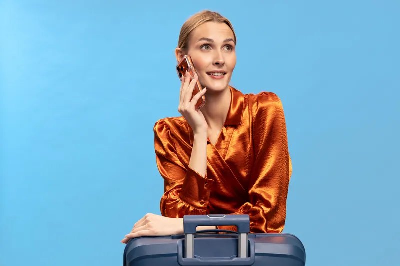 Young woman in orange blouse talking on smartphone while leaning on suitcase handle. Blue background, travel concept with smiling female traveler ready for trip.