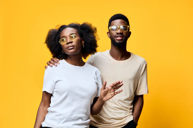Young African couple posing with confidence against a vibrant yellow background, showcasing trendy fashion and stylish sunglasses, embodying modern friendship and joy