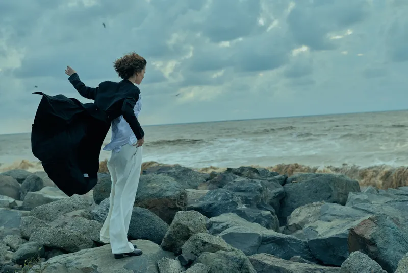 Businessman on rocky shore facing strong wind, coat billowing, contemplating a bold move against the sea and a stormy sky, waves crashing nearby.