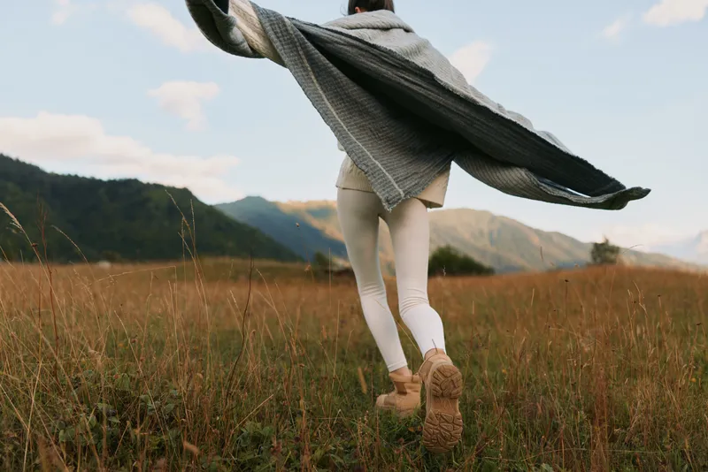 Young woman running across a sunlit field with a flowing cape, embracing outdoor adventure and carefree movement. Natural scenery suggests vitality, wellness, and solo exploration.
