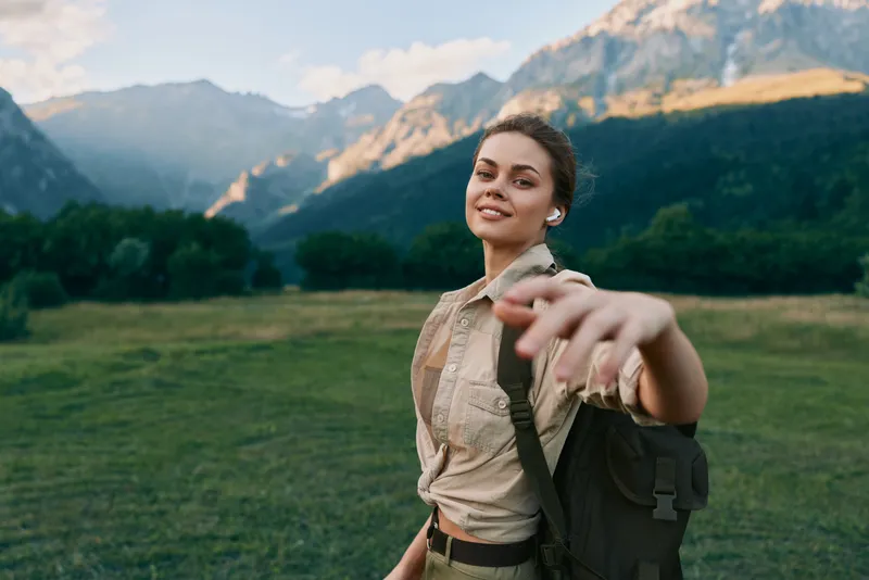 Smiling woman with backpack in a green meadow reaching toward the camera, mountain scenery in the background, outdoor lifestyle, adventure, wellness, nature, portrait.
