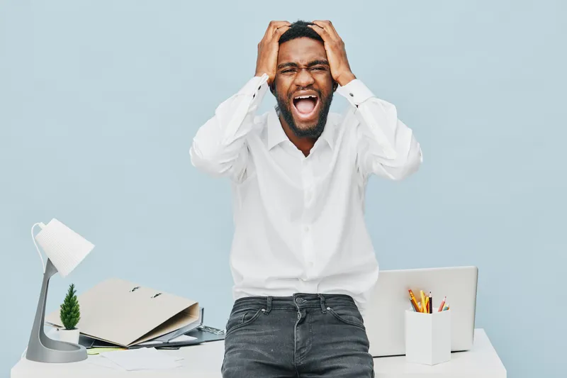 Frustrated young African-American man expressing stress frustration in office environment, holding head with hands, sitting at cluttered desk with open folder, laptop, and stationery, isolated on ligh