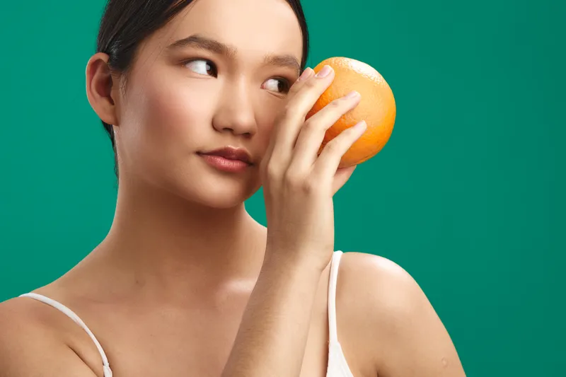 Asian woman holds an orange near her eye against a vibrant green backdrop, examining it with curiosity and calm, creating a fresh, playful studio portrait that feels bright and inviting.