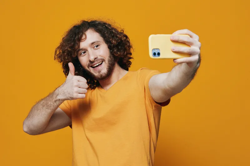 Cheerful young man taking a selfie, wearing a bright yellow t shirt, on a warm orange background, expressing happiness and positivity