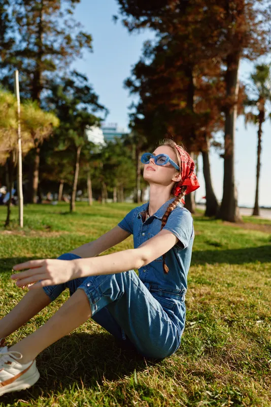 Lifestyle woman in modern boho-western denim street style sitting on grass with sunglasses and red bandana, captured in warm film color tones outdoors.