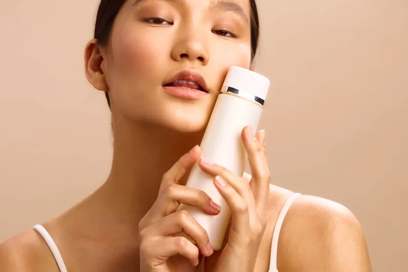 Asian woman gently holds a cosmetic bottle near her face in a soft studio setting, presenting a skincare routine with calm confidence and subtle beauty.