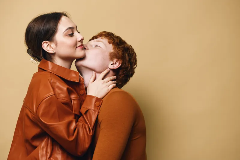 Couple in love embracing indoors, woman in leather jacket kissing man with red hair on cheek, warm clothing and affectionate moment on beige background.