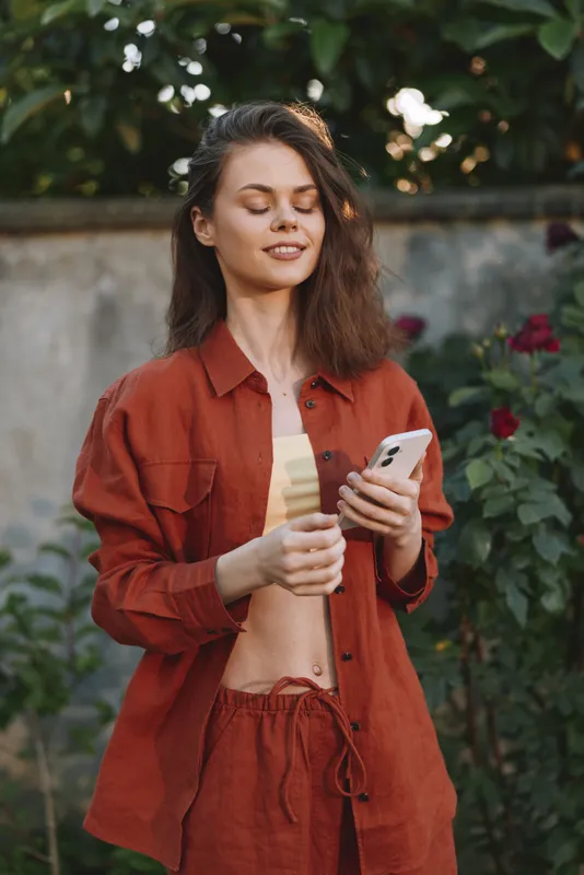 Woman lifestyle portrait, outdoor setting with a smiling woman in red casualwear, holding a phone, enjoying a sunny garden moment